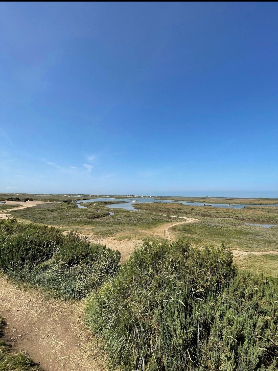 Salt marsh at Brancaster Staithe with tidal creeks, moored boats and a sandy path in the foreground