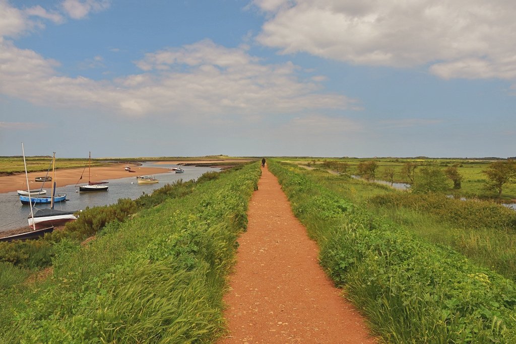 A raised earth path running dead straight through green saltmarsh at Burnham Overy Staithe, with small boats moored in the creek on the left