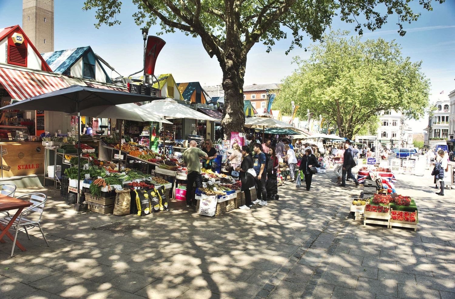Norwich Market stalls under their striped and coloured roofs, shoppers browsing fruit and vegetables in dappled sunlight