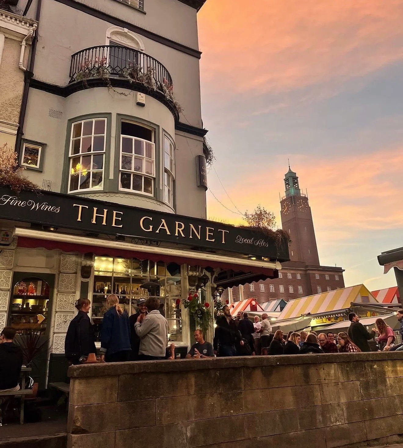 The Garnet on the corner of Norwich market at sunset, drinkers outside with City Hall clock tower behind