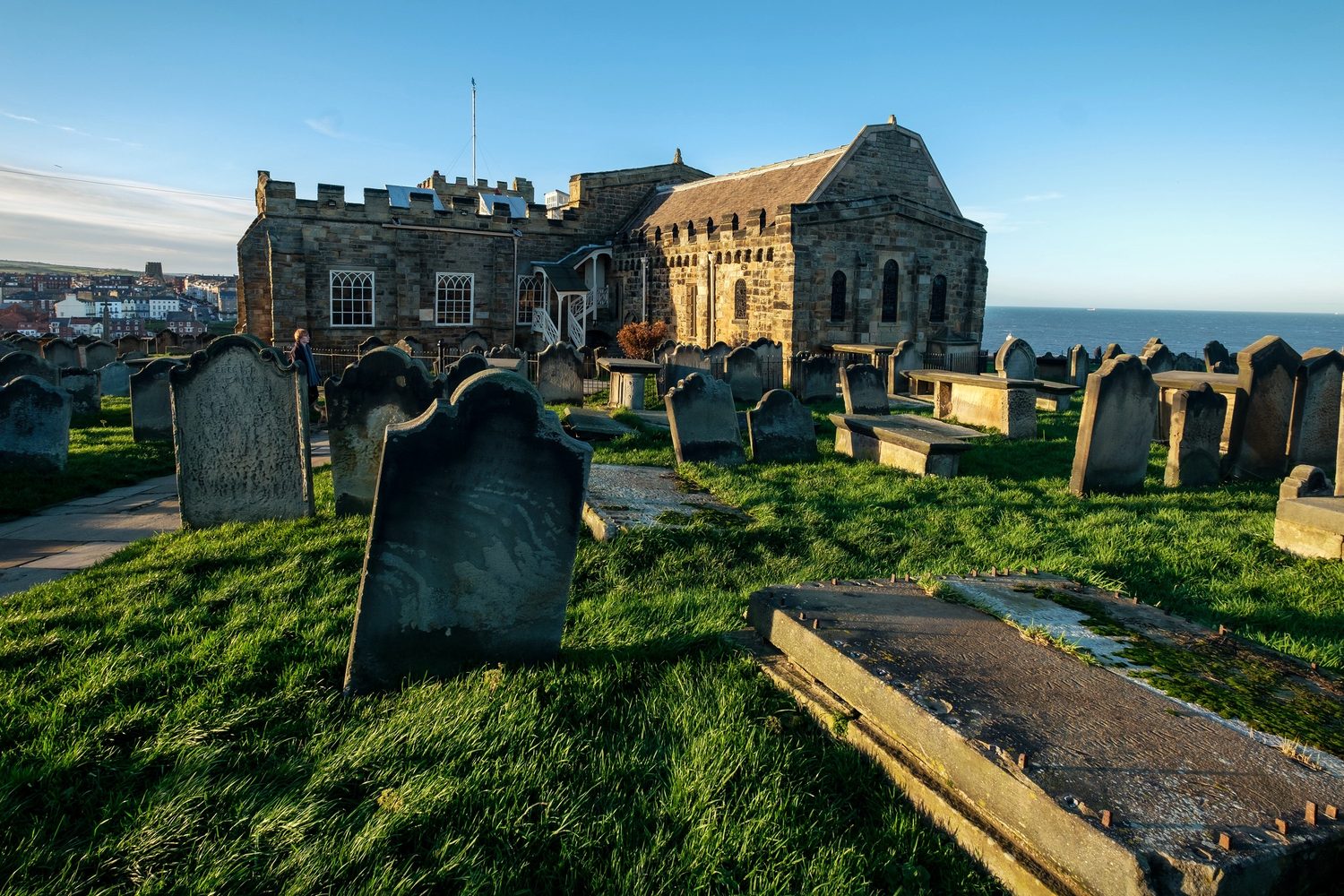 Gravestones in St Mary's churchyard at Whitby with the church behind and the sea to one side, in low golden light