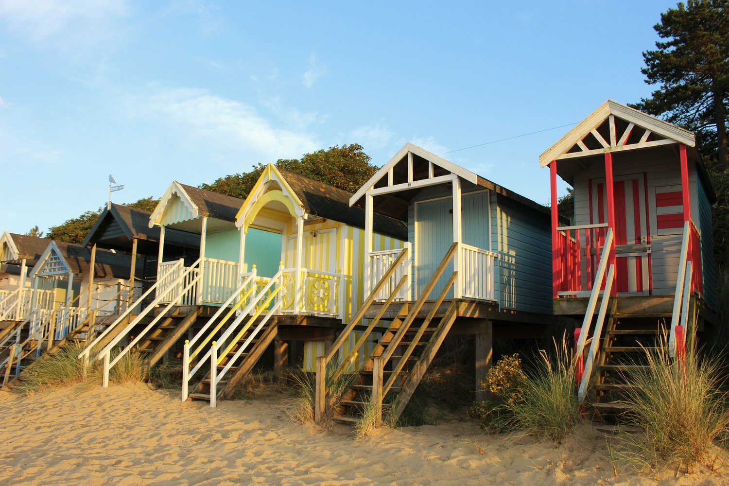A row of brightly painted beach huts on stilts above the sand at Wells-next-the-Sea, with wooden steps and pine trees behind