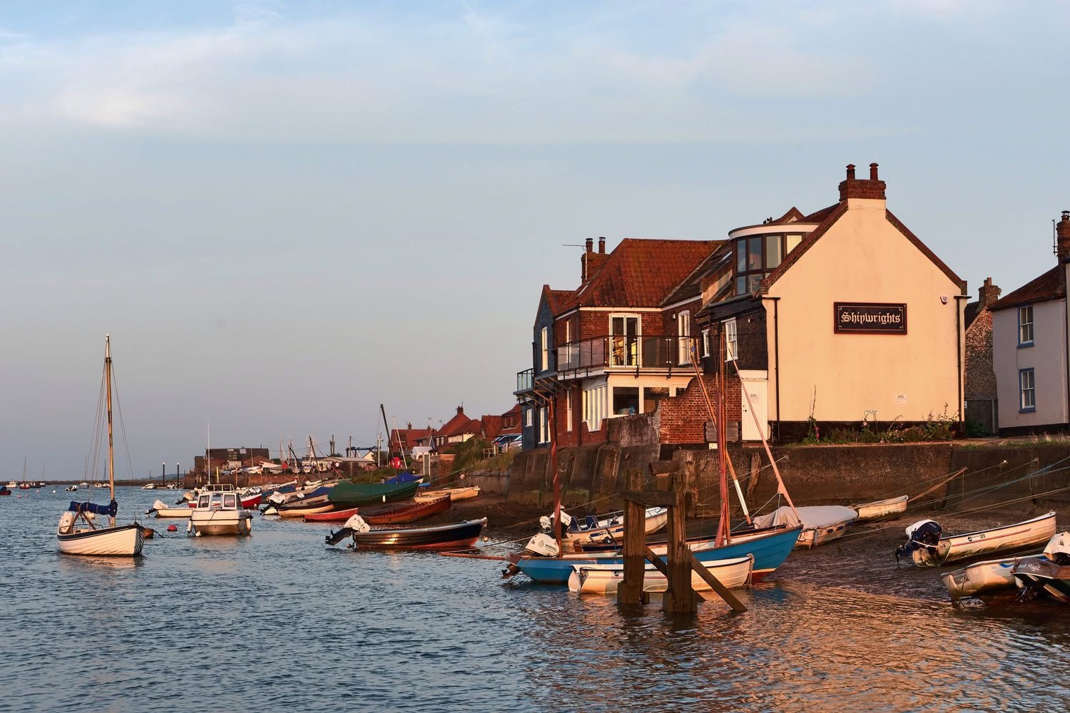 Fishing boats moored along the channel at Wells-next-the-Sea quay in low evening light, with brick and flint houses along the harbour wall
